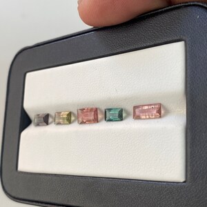 May include: A black leatherette box with a white interior, displaying six rectangular gemstones in various colors, including pink, green, and black.