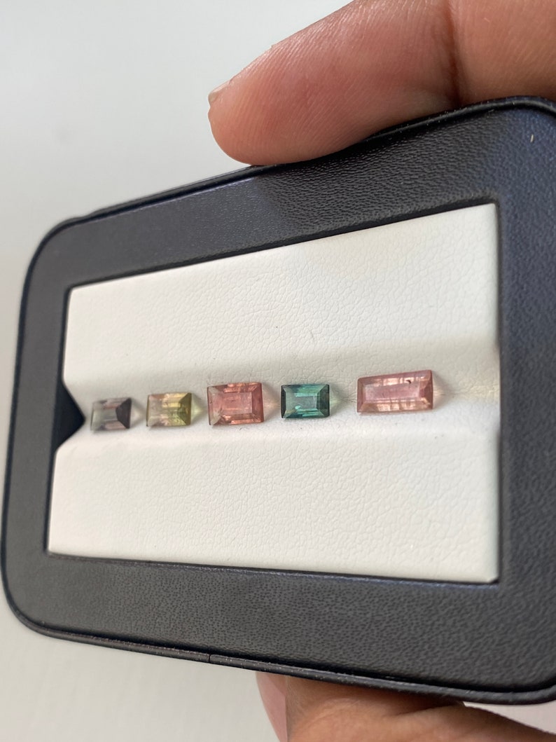 May include: A black leatherette box with a white interior, displaying six rectangular gemstones in various colors, including pink, green, and black.
