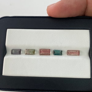 May include: Five rectangular gemstones in a black and white display case. The gemstones are various shades of green, pink, and gray.