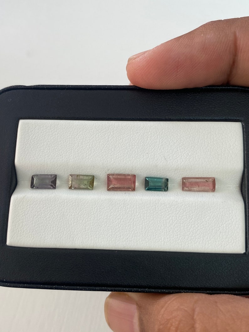 May include: Five rectangular gemstones in a black and white display case. The gemstones are various shades of green, pink, and gray.