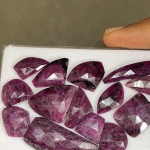 May include: A collection of faceted, deep magenta-colored gemstones with irregular geometric shapes. The stones exhibit a translucent quality with visible internal inclusions and sharp, angular edges, arranged on a white rectangular display tray.