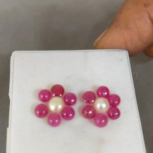 May include: Two pairs of flower-shaped earrings. Each earring features a central white pearl surrounded by six pink gemstones. The earrings are displayed on a white surface.