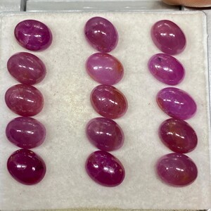 Op de afbeelding: Een verzameling ovaalvormige, gepolijste edelstenen in verschillende tinten diep roze en rood. De edelstenen worden gepresenteerd in een witte tray, waarbij hun gladde, reflecterende oppervlakken worden getoond. De edelstenen zijn waarschijnlijk robijnen, perfect voor sieraden.