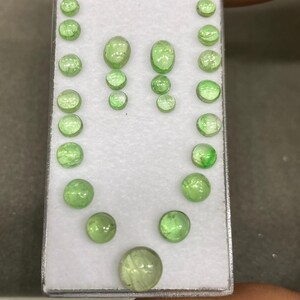 May include: A collection of various-sized, light green gemstones arranged on a white surface within a rectangular container. The gemstones are round, oval, and teardrop-shaped, showcasing a range of sizes and cuts. The stones have a translucent appearance.