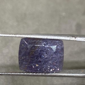 May include: A square-shaped, purple gemstone with a shimmery, iridescent finish. The gemstone is held in tweezers.