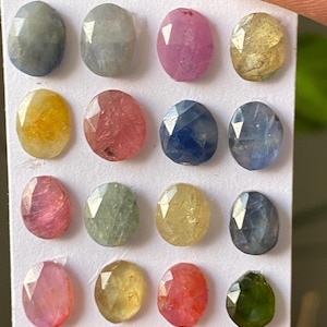 Pode incluir: Uma coleção de dezasseis pedras preciosas facetadas em várias cores, incluindo azul, rosa, amarelo e verde. As pedras ovais estão dispostas num cartão branco, mostrando as suas superfícies polidas e clareza interna. As pedras preciosas são seguradas pelos dedos de uma pessoa.