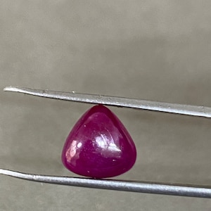 Op de afbeelding: Een gepolijste, dieprode edelsteen, vastgehouden door een pincet. De edelsteen heeft een gladde, afgeronde driehoekige vorm en reflecteert licht. De kleur is een rijk, verzadigd rood, met een lichte doorschijnendheid. De achtergrond is neutraal grijs.