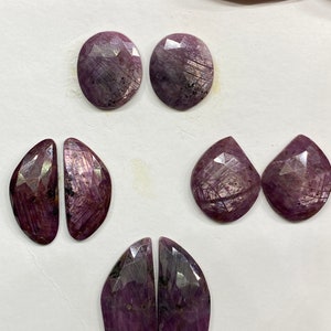Puede incluir: Cinco piedras preciosas púrpuras con superficies facetadas. Las piedras preciosas están dispuestas en pares sobre una superficie blanca.