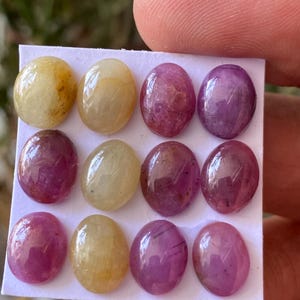 May include: A collection of twelve oval-shaped gemstones in varying shades of yellow and purple. The gemstones are arranged on a white card and held by a person's fingers. The gems have a polished, smooth surface.