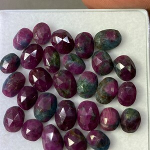 Könnte beinhalten: Eine Sammlung facettierter Rubin-Edelsteine in verschiedenen ovalen und abgerundeten Formen. Die Rubine zeigen eine tiefrote Farbe mit blauen und grünen Farbtönen. Sie sind auf einer weißen Oberfläche angeordnet.