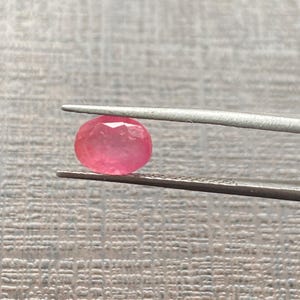 Puede incluir: Una pequeña piedra preciosa rosa de forma ovalada y facetada, sostenida entre las puntas metálicas de unas pinzas. La piedra muestra un tono rosa vibrante y translúcido con múltiples superficies talladas que reflejan la luz.