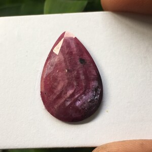 May include: A teardrop-shaped, faceted gemstone with a deep pink and purple hue. The gemstone has a striated pattern and a polished finish.