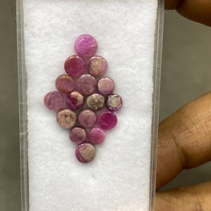May include: A collection of small, round, pink gemstones arranged in a diamond shape within a clear plastic case. The gemstones vary in shade from light pink to deep magenta, with some showing a marbled appearance.