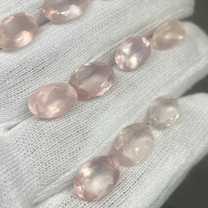 Könnte beinhalten: Eine Sammlung oval geschliffener Rosenquarz-Edelsteine. Die Steine variieren leicht in der Größe und haben eine zarte rosa Farbe. Die Edelsteine werden auf einem weißen, strukturierten Stoff präsentiert, der ihre polierten Oberflächen zeigt.