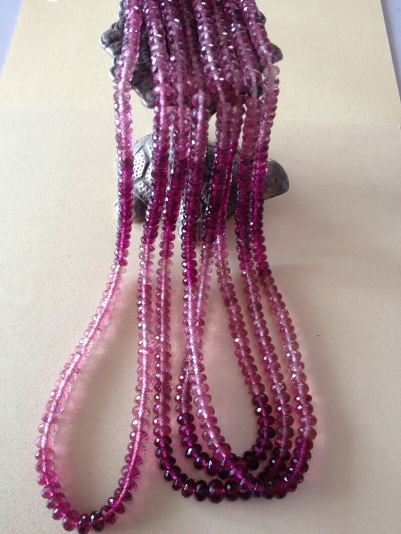 May include: Multiple strands of pink and purple faceted beads. The beads are strung together, resembling necklaces. The colour gradient ranges from light pink to deep purple. The beads are likely made of a translucent material, catching the light.