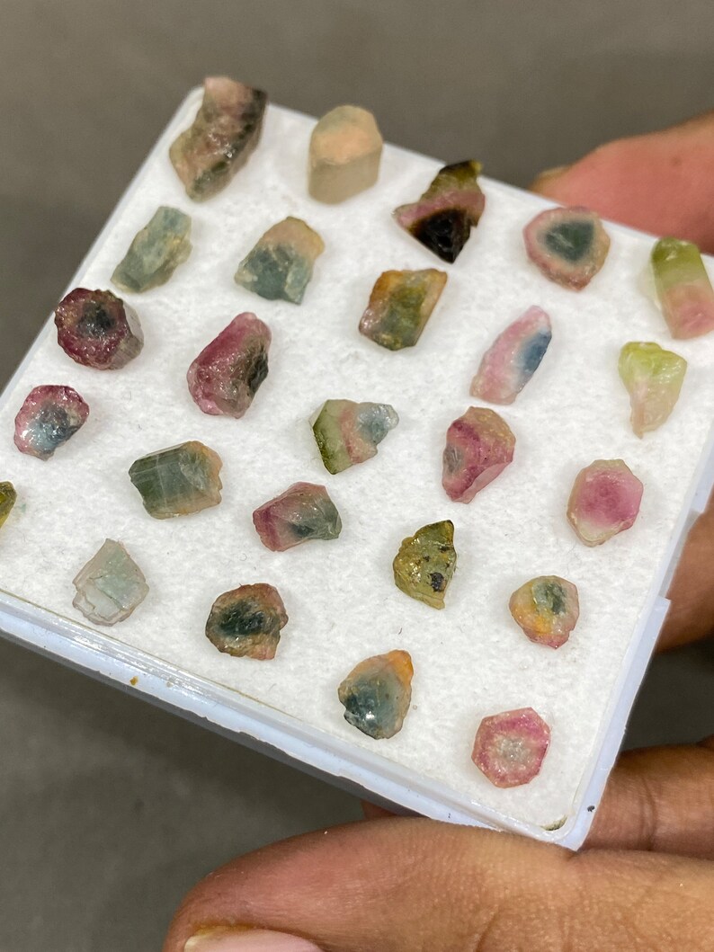 May include: A collection of 24 small, colourful, raw tourmaline crystals in a white foam-lined box. The crystals are various shades of pink, green, blue, and black.