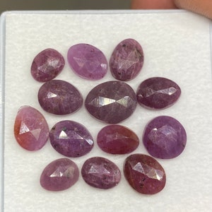 Peut inclure: Une collection de onze pierres précieuses ovales à facettes, de couleur rose et violette, disposées sur une surface blanche. Chaque pierre présente une surface polie à facettes multiples qui reflète la lumière, révélant des nuances de magenta.