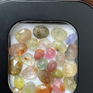 Pode incluir: Uma coleção de pedras preciosas facetadas em uma variedade de cores, incluindo amarelo, laranja, rosa e roxo. As pedras preciosas estão contidas em um pequeno recipiente quadrado preto. As pedras preciosas são de vários tamanhos e formas.
