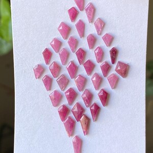 Peut inclure: Collection de petites pierres précieuses roses facettées disposées sur une surface blanche. Les pierres précieuses ont une forme de losange avec des extrémités pointues et des nuances de rose variées. L'arrangement forme un losange.