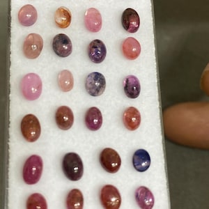 May include: A selection of oval gemstones in shades of pink, brown, and purple. The gemstones are presented in a white tray, highlighting their polished surfaces and natural variations. The gemstones are of different sizes.