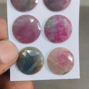 Puede incluir: Seis piedras preciosas redondas y facetadas dispuestas en dos columnas sobre una tarjeta blanca. Las piedras muestran colores variados que incluyen violeta intenso, magenta, rosa, azul y melocotón claro, cada una con una superficie tallada.