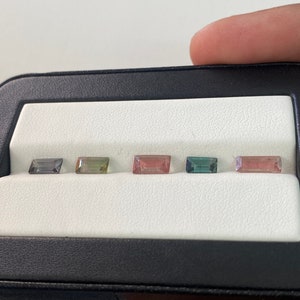 May include: Five rectangular cut gemstones in a black and white display case. The gemstones are a variety of colors, including green, pink, and blue.