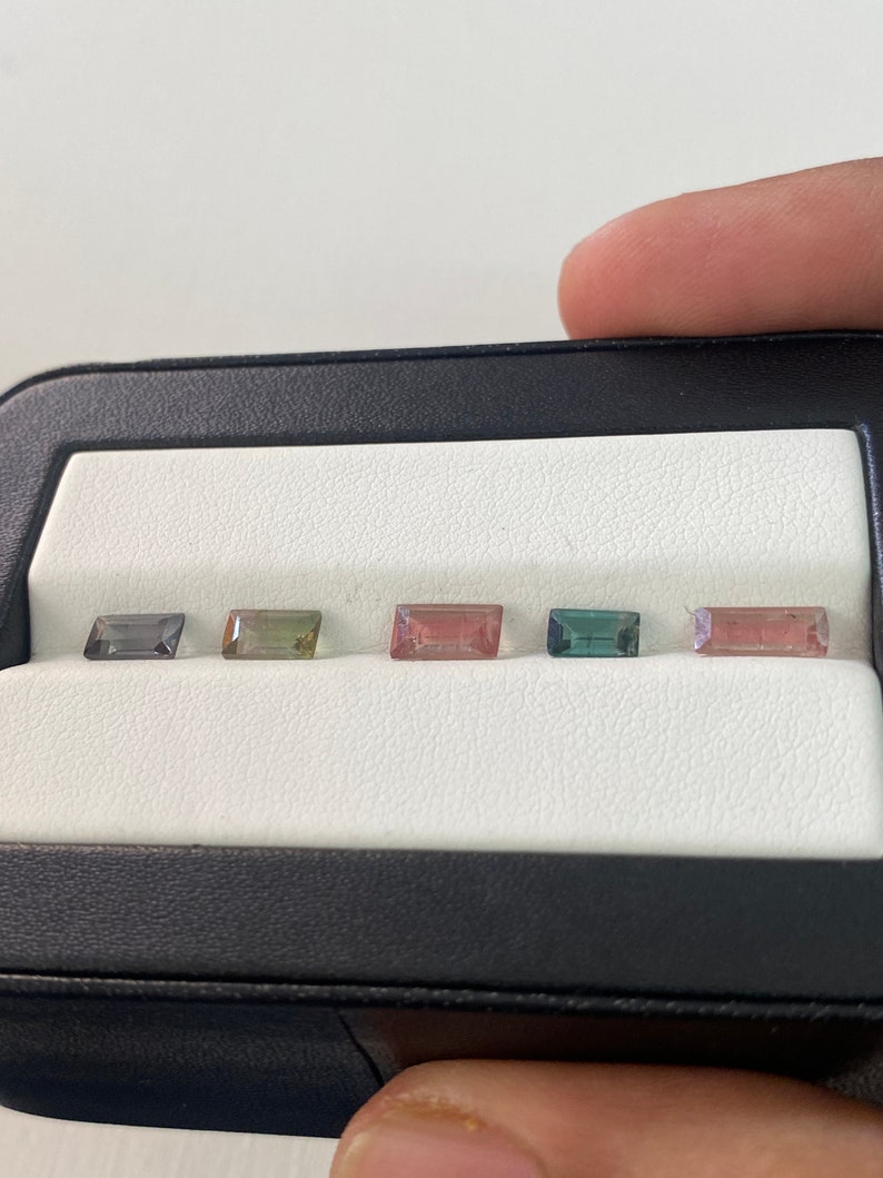 May include: Five rectangular cut gemstones in a black and white display case. The gemstones are a variety of colors, including green, pink, and blue.