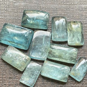 May include: A collection of rectangular, translucent aquamarine gemstones. The stones vary in shades of blue and green, with visible internal textures and inclusions. The gemstones are arranged on a textured surface, showcasing their polished surfaces and natural variations.
