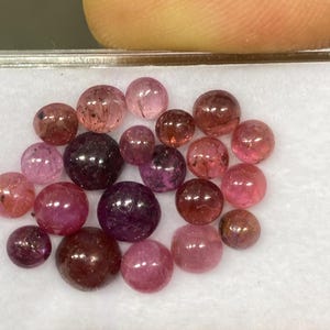 May include: A collection of polished, round gemstones in shades of pink, red, and purple. The gemstones are displayed in a clear, rectangular container. The stones vary in size and color saturation, with some appearing translucent.