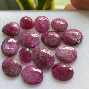 May include: A collection of twelve pink ruby gemstones, each with a faceted oval shape. The gemstones are displayed on a white surface.
