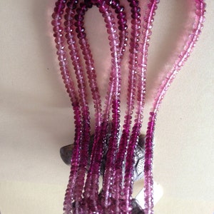 May include: A collection of pink and purple beaded necklaces. The beads are round and faceted, with a colour gradient from deep purple to pale pink. The necklaces are displayed against a neutral background.