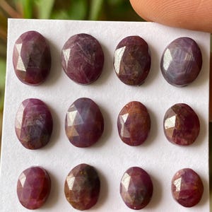 Può includere: Una collezione di dodici rubini sfaccettati di forma ovale disposti in tre file su un cartoncino bianco. Ogni gemma presenta una tonalità dal rosso intenso al rosso violaceo con inclusioni interne visibili e una superficie lucida e sfaccettata.