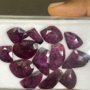 Puede incluir: Una colección de doce rubíes facetados de color magenta intenso dispuestos en una caja de presentación de plástico transparente. Cada piedra preciosa presenta una forma geométrica irregular única con superficies pulidas que reflejan la luz.