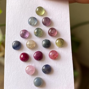 May include: A collection of small, round gemstones in various colors, including pink, red, blue, green, and yellow. The gemstones are displayed on a white card, showcasing their polished surfaces and vibrant hues. These gemstones are perfect for jewelry making.
