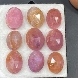 May include: Nine oval gemstones in pink, peach, and orange hues are displayed on a white surface. Each stone features a faceted cut, creating multiple reflective surfaces. The gemstones vary in colour intensity and clarity.