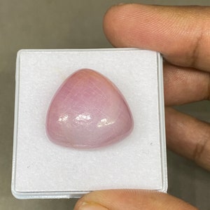 Könnte beinhalten: Ein polierter, dreieckiger Edelstein in einem zarten Rosa. Der Edelstein wird in einem weißen Kunststoffgehäuse präsentiert, das seine glatte, reflektierende Oberfläche hervorhebt. Die Form des Steins ist ein abgerundetes Dreieck mit subtilen inneren Mustern.