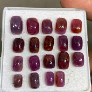 May include: A collection of 18 faceted, rectangular, purple and red gemstones in a white foam-lined box.
