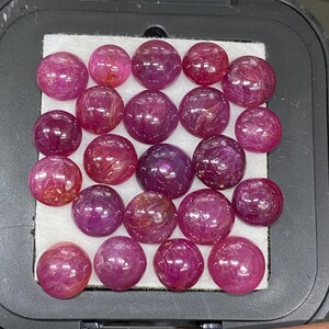 May include: A collection of nineteen round, cabochon-cut rubies arranged in a grid pattern inside a black display case. The gemstones exhibit varying shades of deep pink and red with visible internal inclusions and a smooth, polished surface finish.