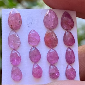 May include: A collection of 12 pink pear-shaped gemstones on a white card. The gemstones are faceted and have a translucent appearance.