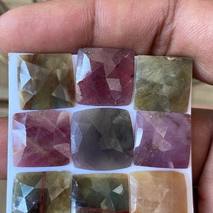 Può includere: Nove gemme quadrate nei toni del viola, rosa e verde oliva sono disposte su una superficie bianca. Ogni pietra ha molteplici sfaccettature, creando un effetto scintillante. Le gemme sono tenute da una mano, suggerendo che si tratti di pietre sciolte.