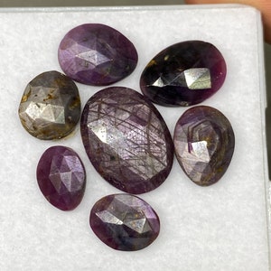 Puede incluir: Seis piedras preciosas ovaladas de color púrpura y plateado en una caja blanca. Las piedras preciosas tienen un corte facetado y están dispuestas en un racimo.
