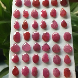 May include: A collection of thirty-six faceted, pear-shaped and oval-shaped pink and red rubies arranged in a grid on a white rectangular display card. Each gemstone features multiple polished surfaces that reflect light, showcasing various shades of pink.