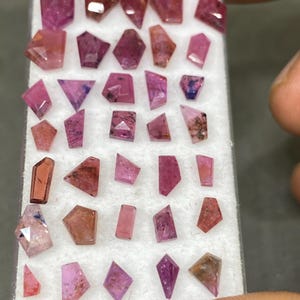 May include: A collection of faceted gemstones in various shapes and shades of pink and red. The gemstones are displayed in a white tray, showcasing their unique cuts and clarity. The assortment includes rectangular, trapezoid, and irregular shapes.