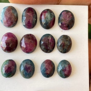 Könnte beinhalten: Eine Auswahl von zwölf ovalförmigen Edelsteinen. Die Edelsteine zeigen eine Mischung aus Farben, darunter tiefes Rot, Blau und Grün, mit einer facettierten Oberfläche. Die Steine sind auf einer weißen Oberfläche angeordnet.
