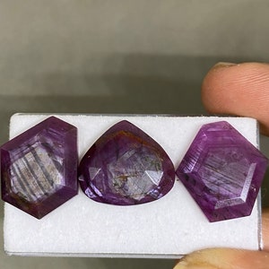 Puede incluir: Tres gemas facetadas en tonos morados y granates se exhiben sobre una superficie blanca. Las gemas tienen formas geométricas únicas, incluyendo un hexágono, una lágrima y otro hexágono. Las gemas tienen estrías internas.
