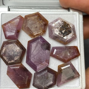 Könnte beinhalten: Eine Sammlung von acht facettierten violetten und braunen Edelsteinen in einer weißen Schachtel. Die Edelsteine sind in verschiedenen Formen und Größen.