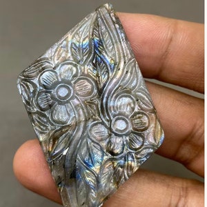 May include: A carved gemstone with a floral design. The stone is diamond-shaped and features detailed carvings of flowers and leaves. The stone has a mix of gray, blue, and iridescent colors. The gemstone is held in a hand.