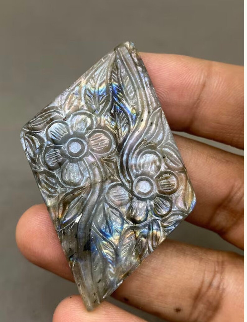 May include: A carved gemstone with a floral design. The stone is diamond-shaped and features detailed carvings of flowers and leaves. The stone has a mix of gray, blue, and iridescent colors. The gemstone is held in a hand.