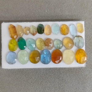 Puede incluir: Una tarjeta de presentación rectangular blanca que contiene veinticuatro piedras preciosas facetadas de diversas formas y colores, incluyendo tonos amarillos, azules, verdes y naranjas. Las piedras están dispuestas en tres filas horizontales.