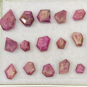 May include: A selection of pink gemstones, cut into various geometric shapes, presented in a white tray. The gems show internal patterns and inclusions, and are arranged in rows. The colours range from light to deep pink.